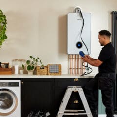 A man in a black uniform stands on a stepladder inspecting a white boiler mounted on a wall in a modern laundry room. The room features a washing machine, plants, and shelves with decorative items. A radiator and coats are on the right.