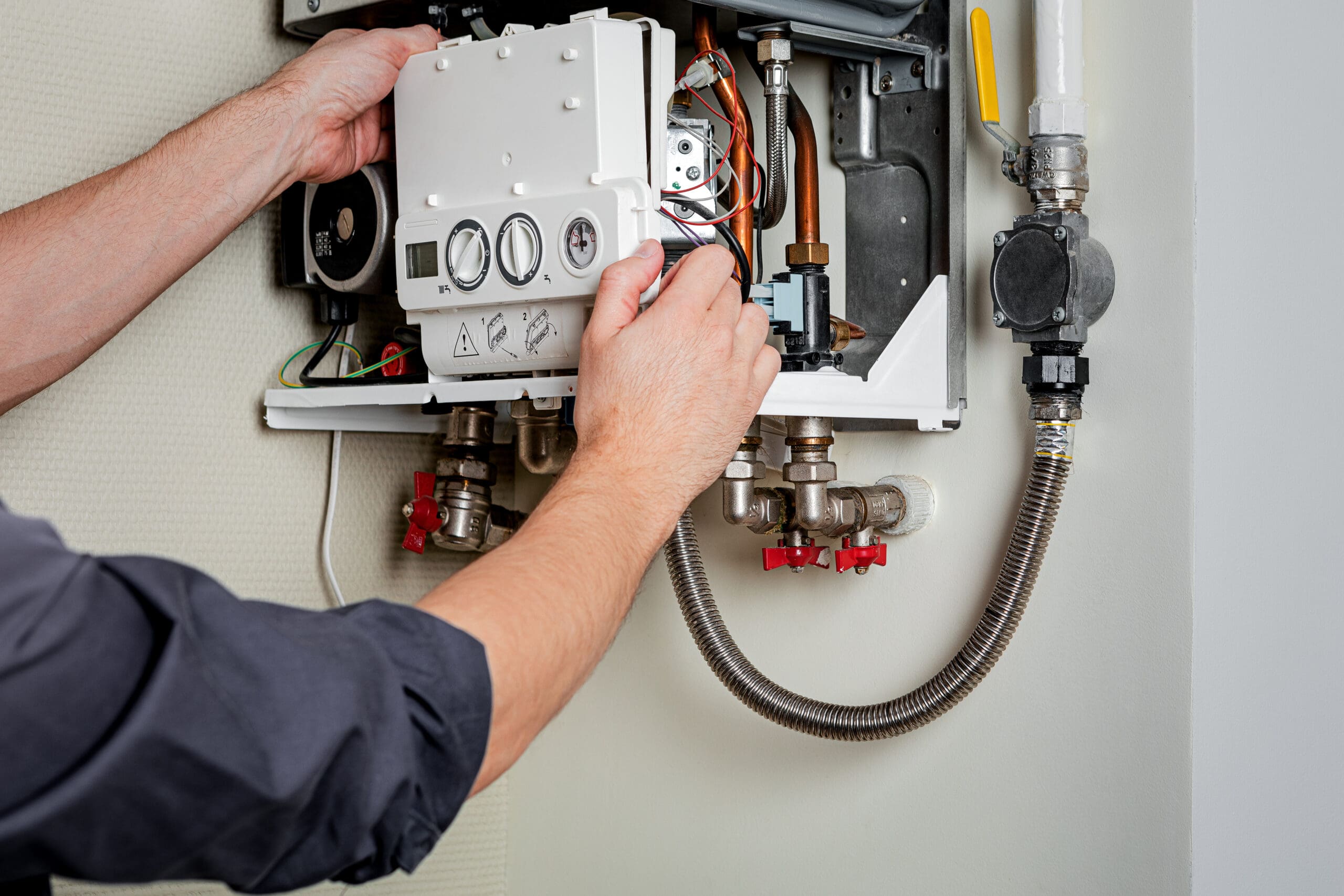 A person adjusts a white control panel on a wall-mounted boiler, surrounded by various pipes and gauges. The person is wearing a dark shirt and is using both hands to handle the equipment.