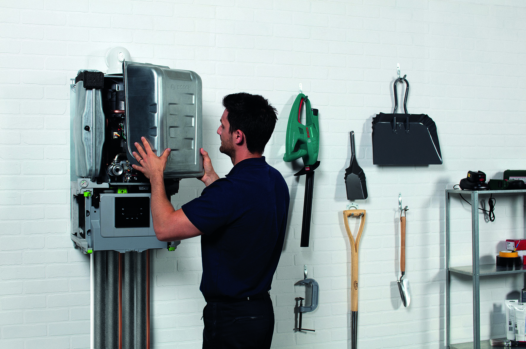 A person is working on a wall-mounted boiler in a garage or workshop. Tools like a shovel, spade, and hedge trimmer are hanging on the wall nearby. Shelving with additional tools is visible in the background.