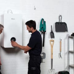 Two men stand by a white boiler mounted on a wall, with one demonstrating its use. The wall also displays various tools and gardening equipment. A shelf with paint cans and tools is visible to the right.