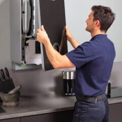 A person wearing a navy blue uniform is installing or maintaining a wall-mounted appliance in a kitchen. The countertop has a blender and a knife block. The appliance cover is partially opened.