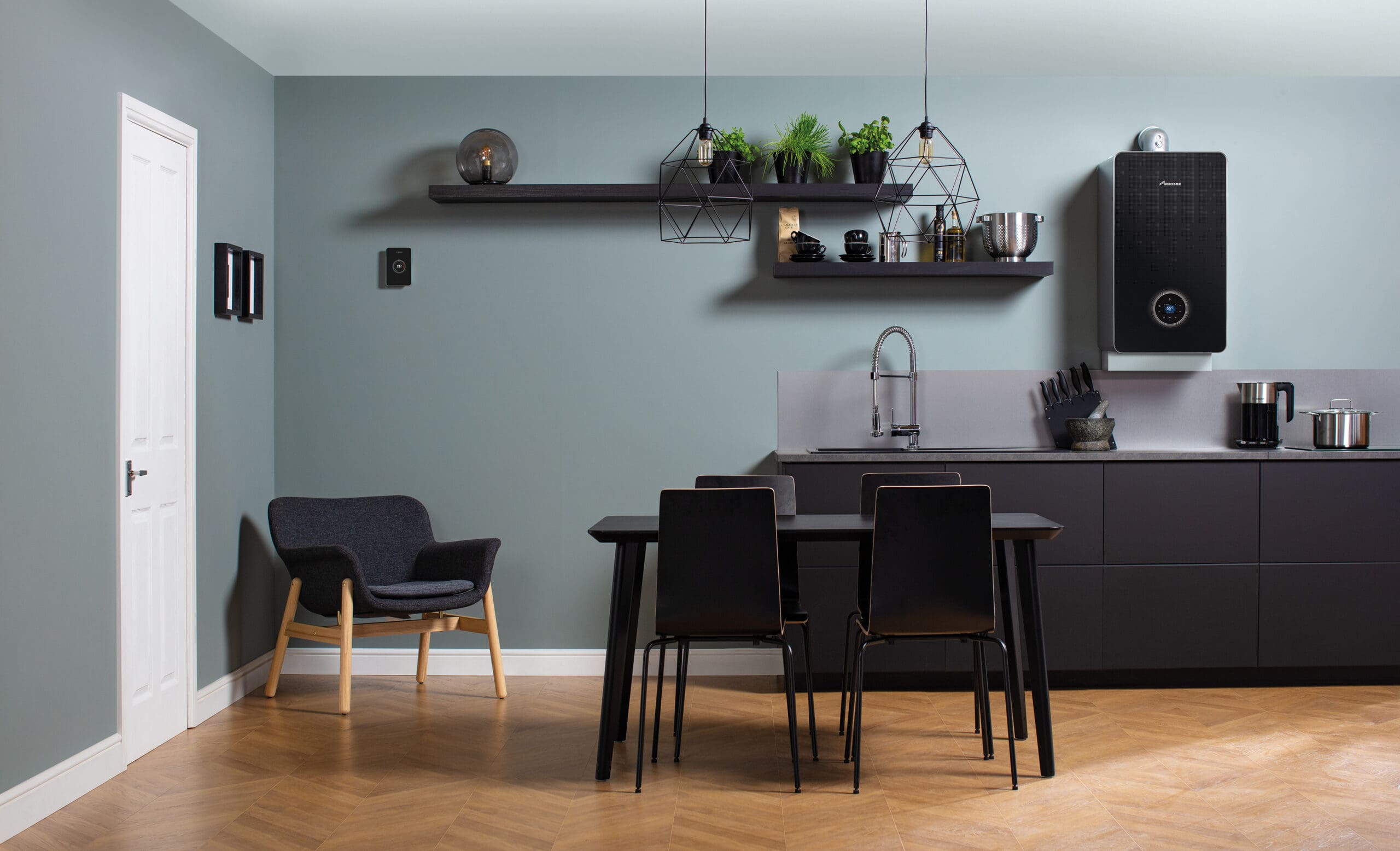 Modern kitchen with light blue walls, dark cabinets, and wood flooring. Features a dining table with chairs, wall-mounted shelves holding plants, and a black boiler on the wall. A dark chair sits in the corner. Minimalistic decor.