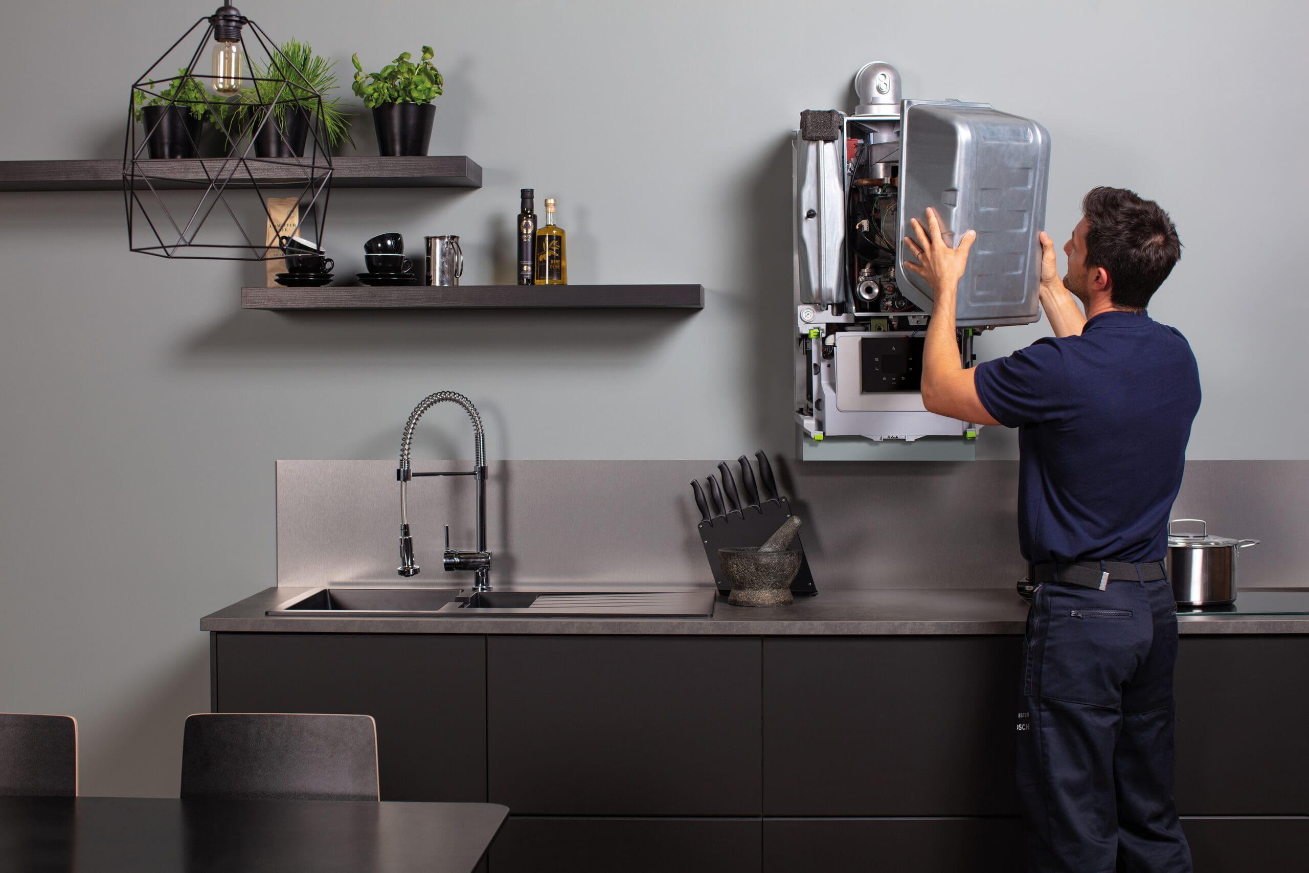 A man in a navy uniform is installing or repairing a wall-mounted appliance in a modern kitchen. The kitchen features a sleek black countertop, a sink, utensils, and potted plants on shelves. A geometric light fixture hangs above.