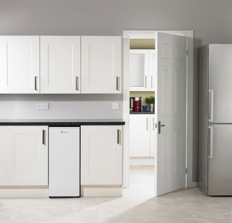 A modern kitchen with white cabinets, a small white fridge, and a silver full-size refrigerator. A door is slightly open, leading to an adjacent room with more cabinets and kitchen items, including a red kettle and green plant. Light tile flooring.