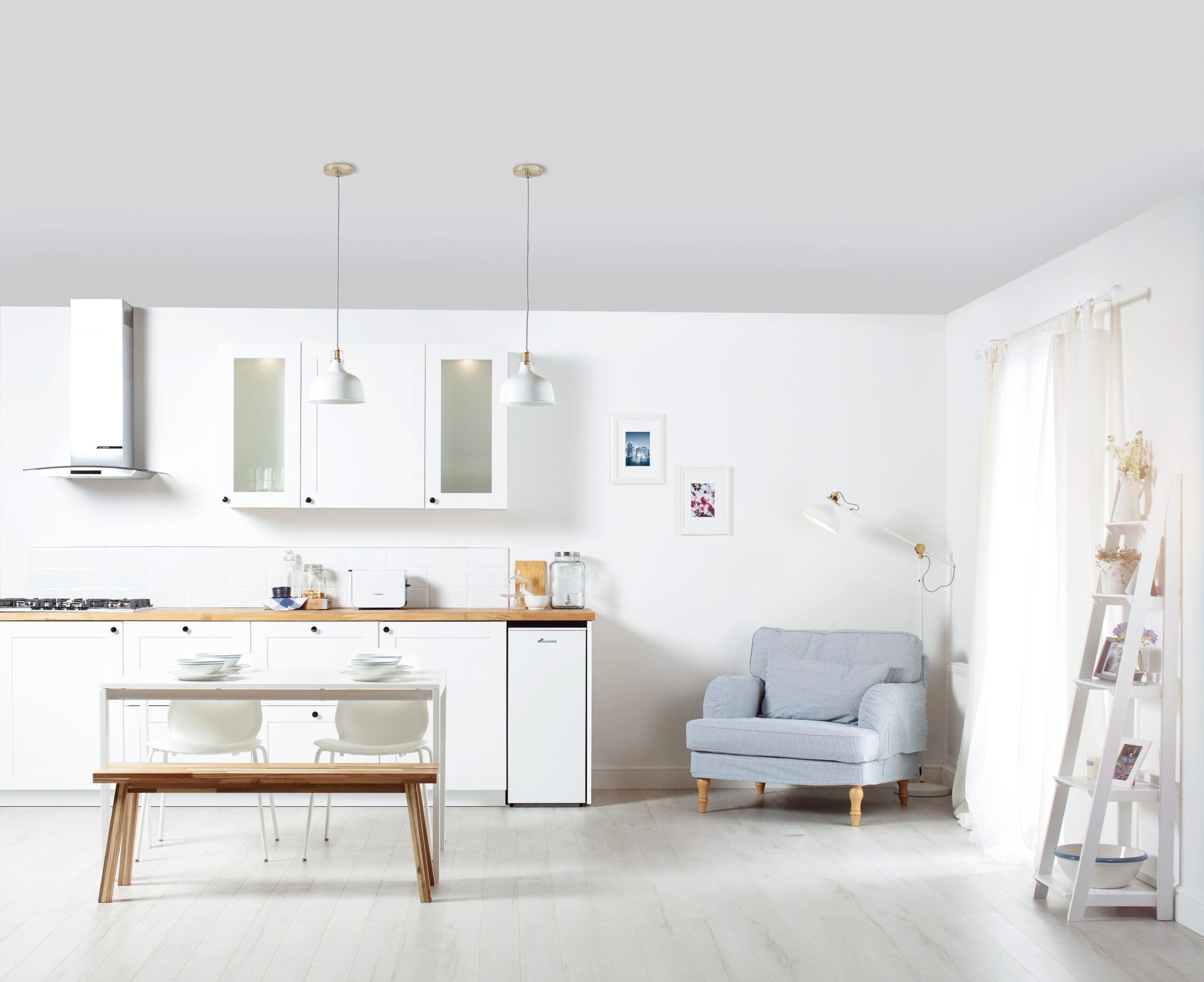 A minimalist kitchen and dining area with white cabinets, a small dining table with a bench and chairs, a light blue armchair, framed pictures on the wall, and a ladder bookshelf. Natural light flows through a large window with sheer curtains.