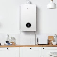 A modern white boiler mounted on a kitchen wall with wooden countertops. On the countertop, there are jars, a toaster, and kitchen utensils. The room is well-lit with a white pendant light.