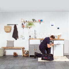 A technician in a navy uniform is kneeling in a modern kitchen, repairing a dishwasher. Tools are on a nearby cloth, and a black tool bag is on the floor. Light gray cabinets and plants are part of the minimalist decor.