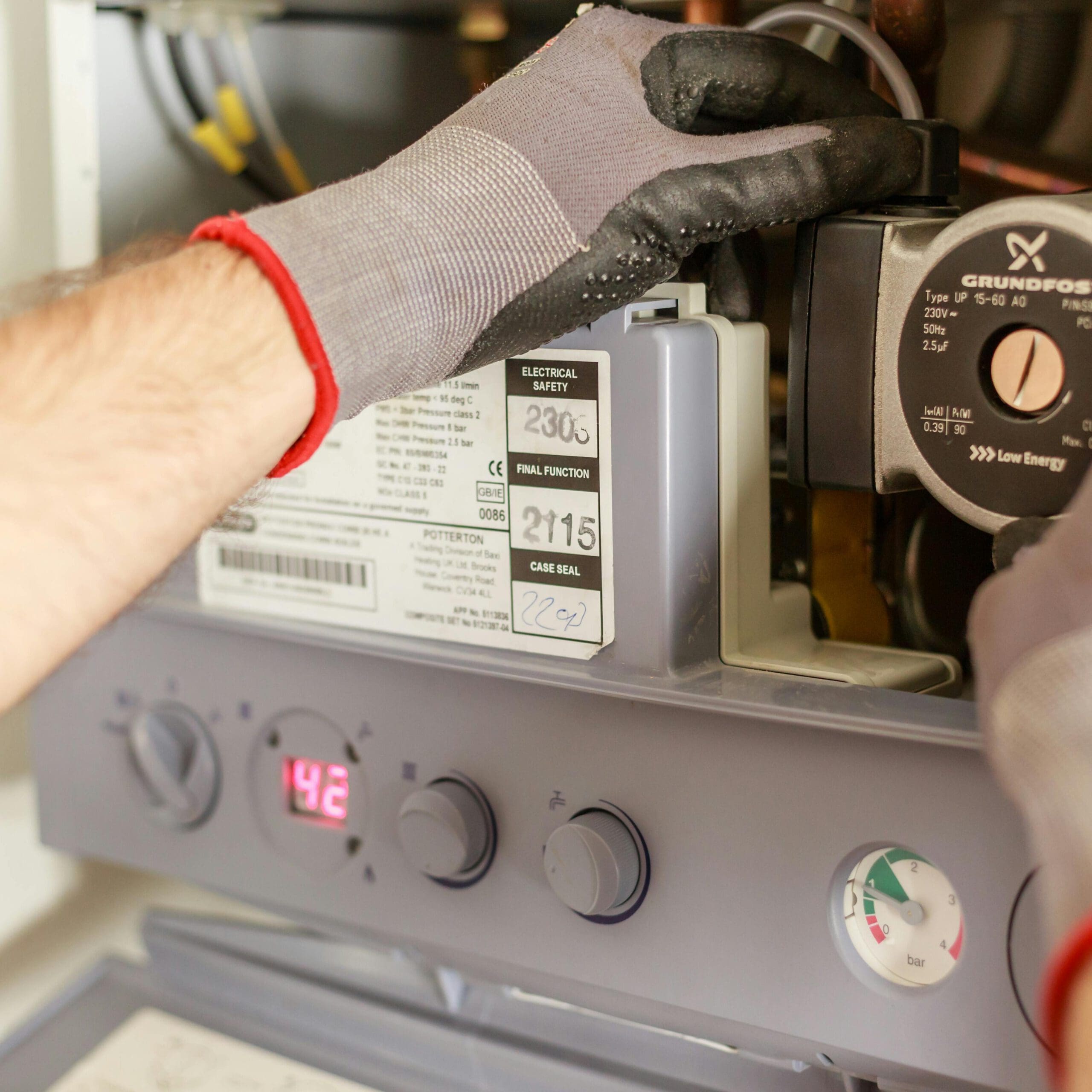 A person wearing gloves adjusts controls on a boiler system. The panel displays a digital reading of 42. The equipment includes a Grundfos water pump. Various dials and labels are visible, indicating settings and specifications.