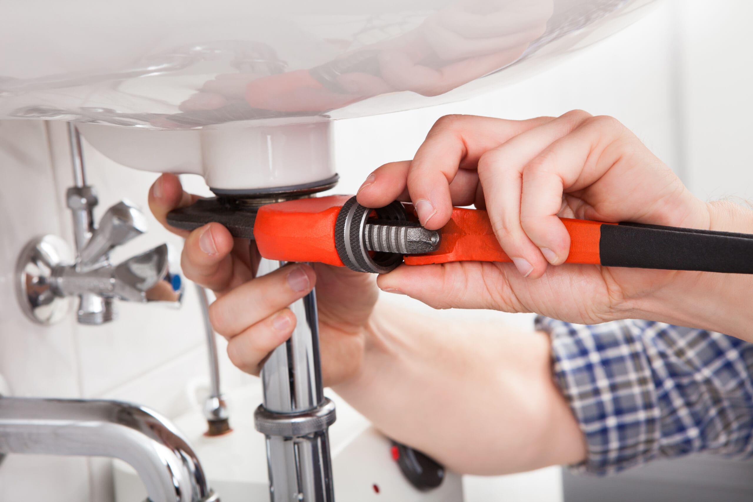 A person wearing a plaid shirt uses an orange pipe wrench to tighten a fitting under a white sink. The scene shows plumbing tools and chrome pipes in a bathroom setting.