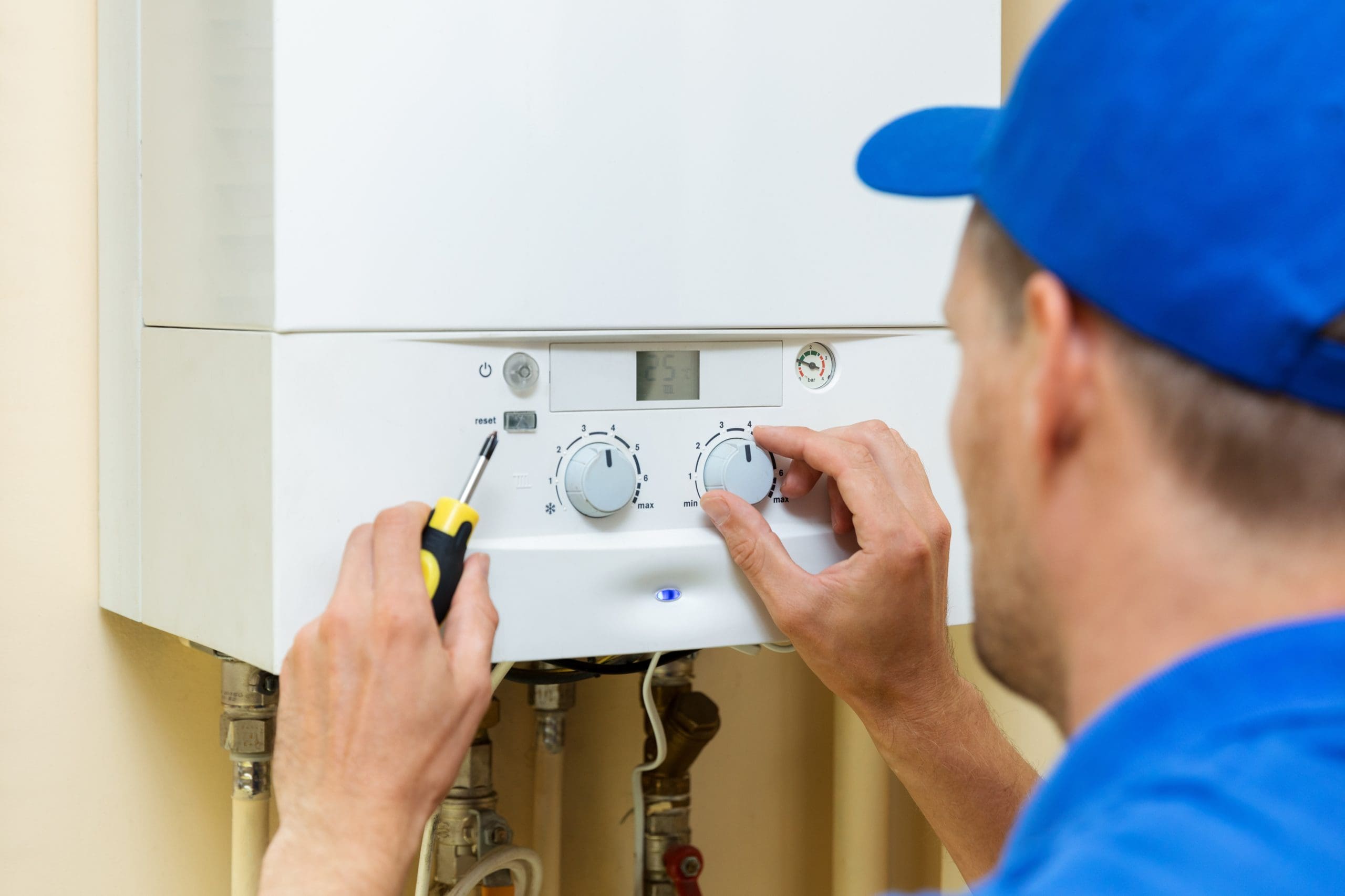 A technician in a blue cap is adjusting the dials on a white boiler, using a screwdriver. The boiler is mounted on a wall and has a small display and several gauges.