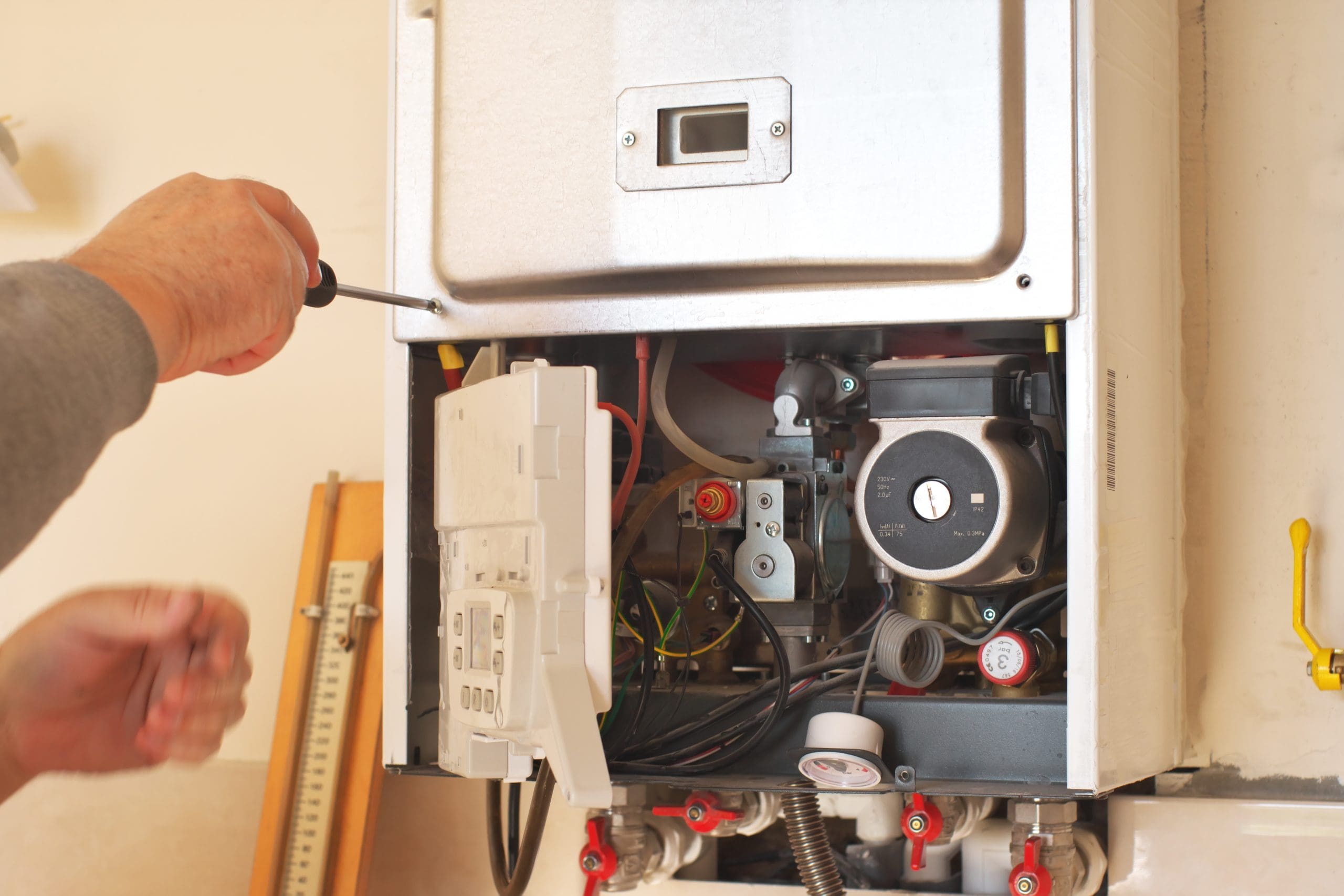 A person using a screwdriver works on the inner components of a wall-mounted boiler. The front panel is open, revealing wiring and mechanical parts.