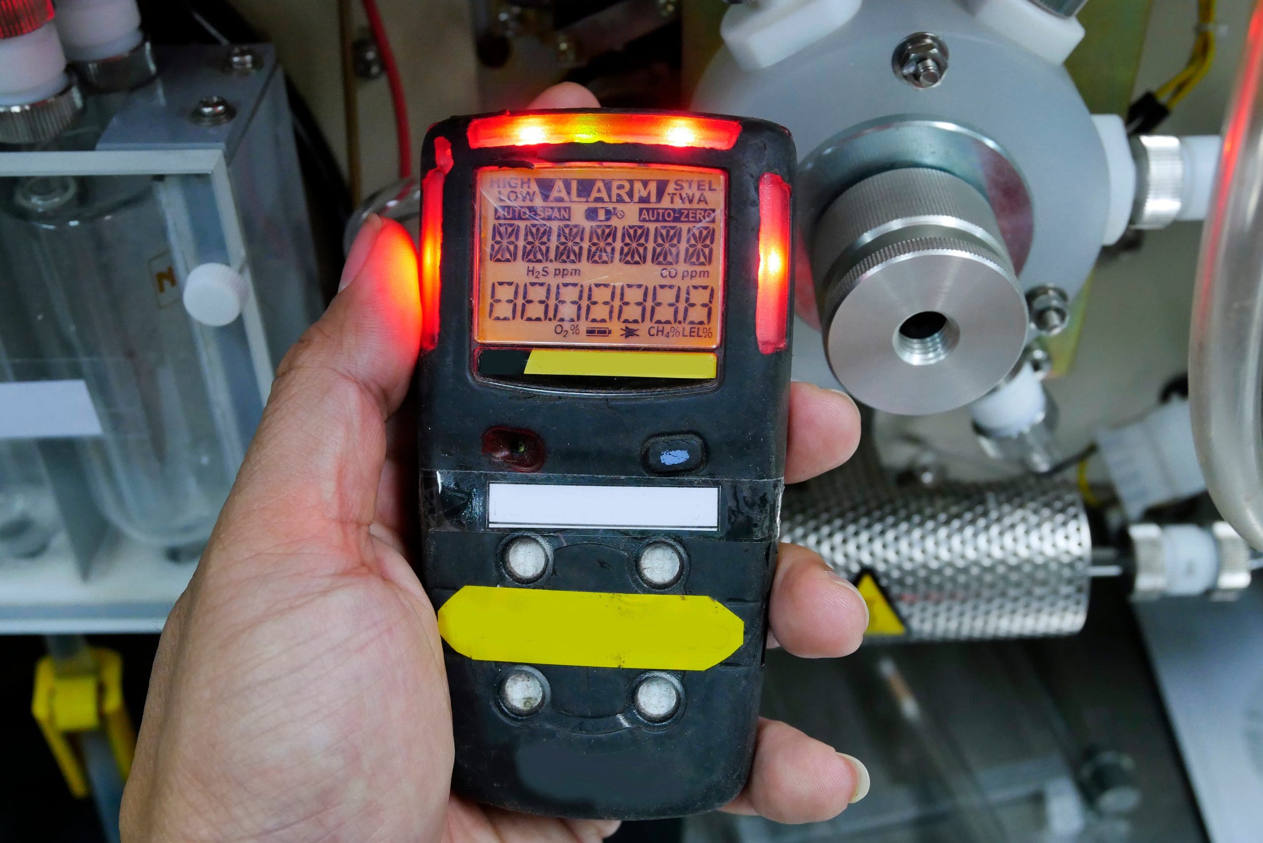 A person holds a gas detector with a digital display and several illuminated LED indicators. The device shows numeric readings and is surrounded by industrial equipment.