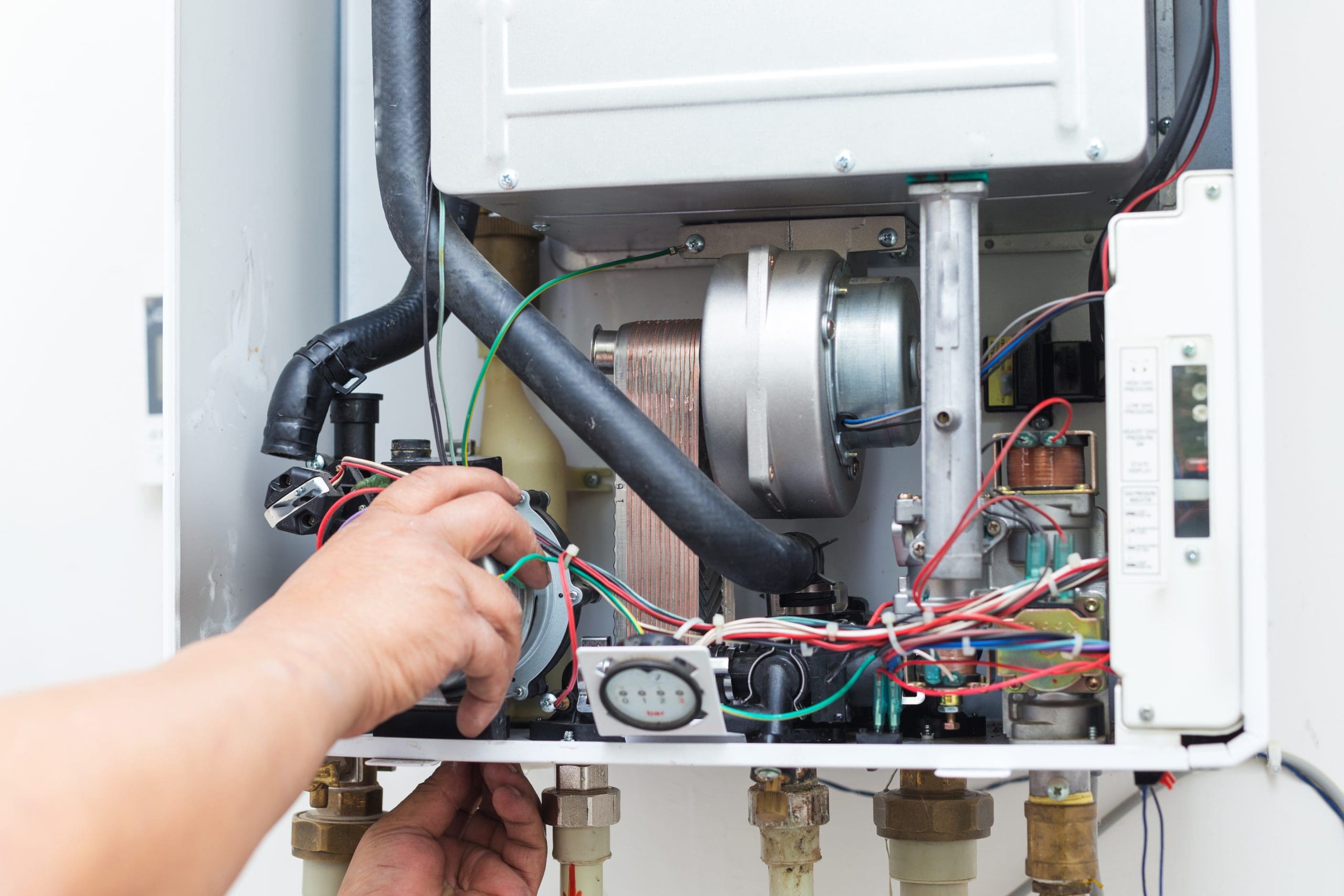 A close-up of a person repairing or inspecting the internal components of a boiler or central heating system. Wires, pipes, and various mechanical parts are visible, along with hands adjusting or holding parts of the machinery.