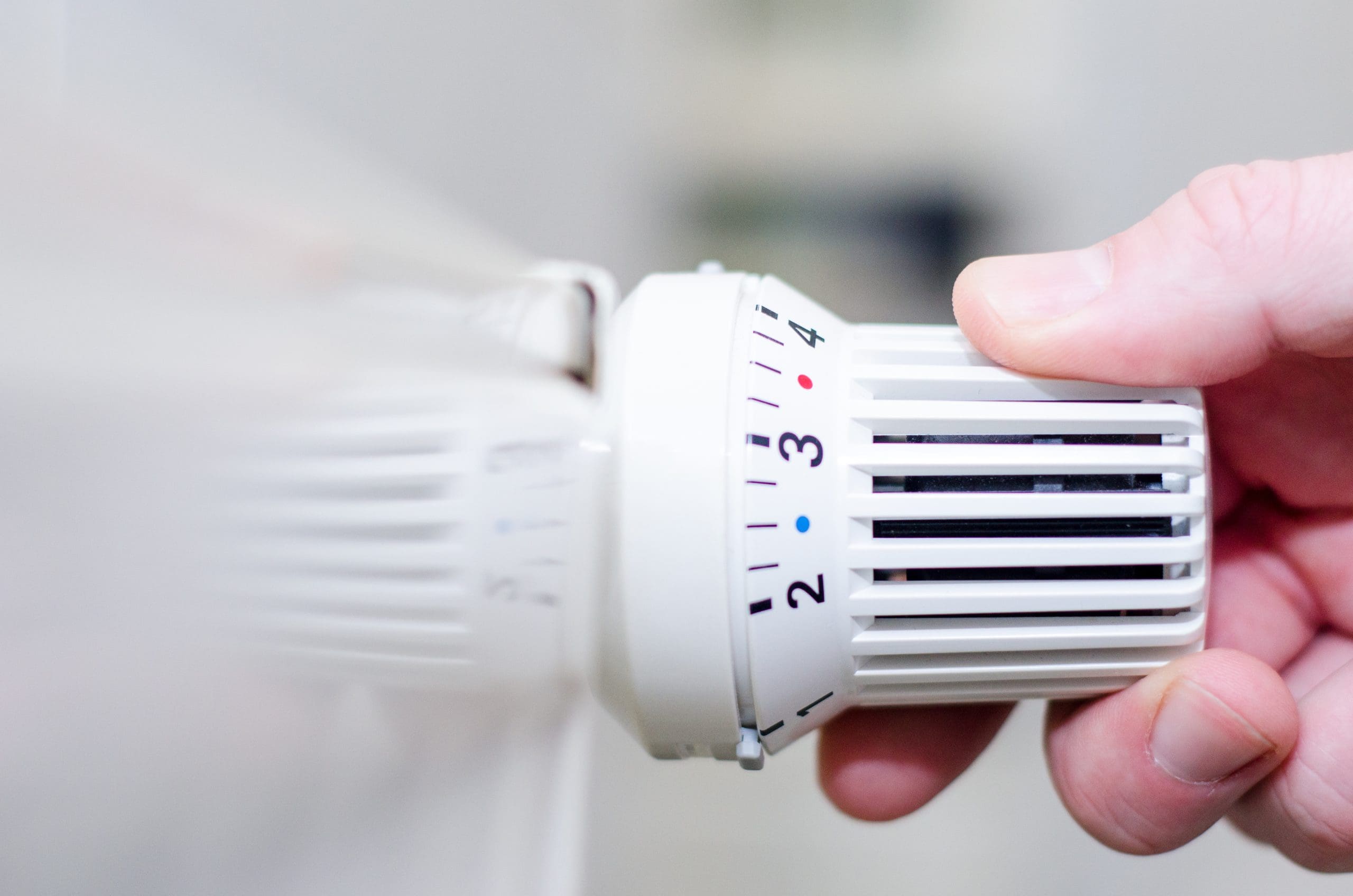 A person adjusts a white radiator thermostat, turning the dial from setting 3 to setting 4.