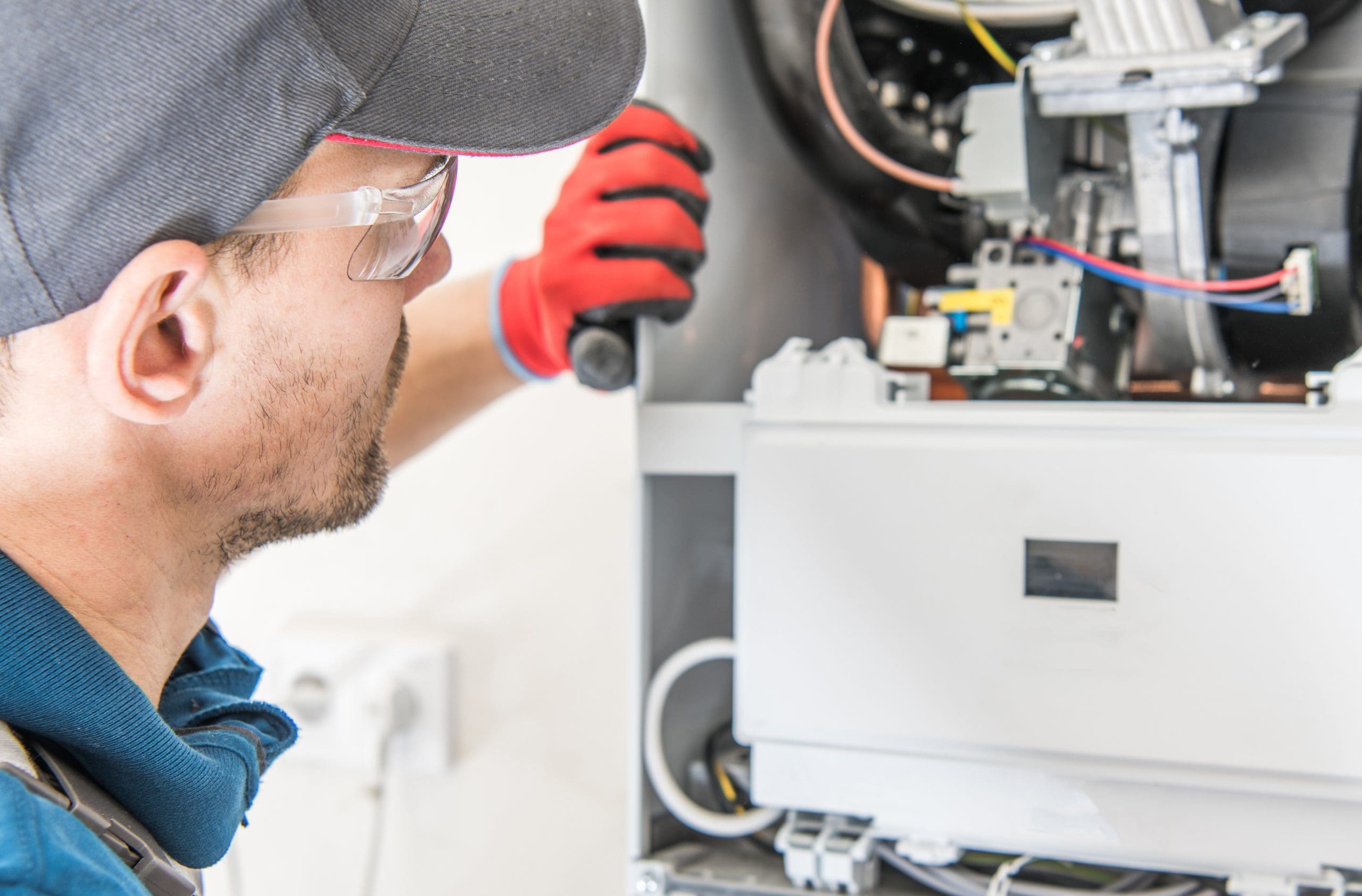 A person wearing a cap, safety glasses, and gloves is inspecting or repairing an HVAC system. They are using a tool and examining the equipment closely. The setting appears to be indoors.