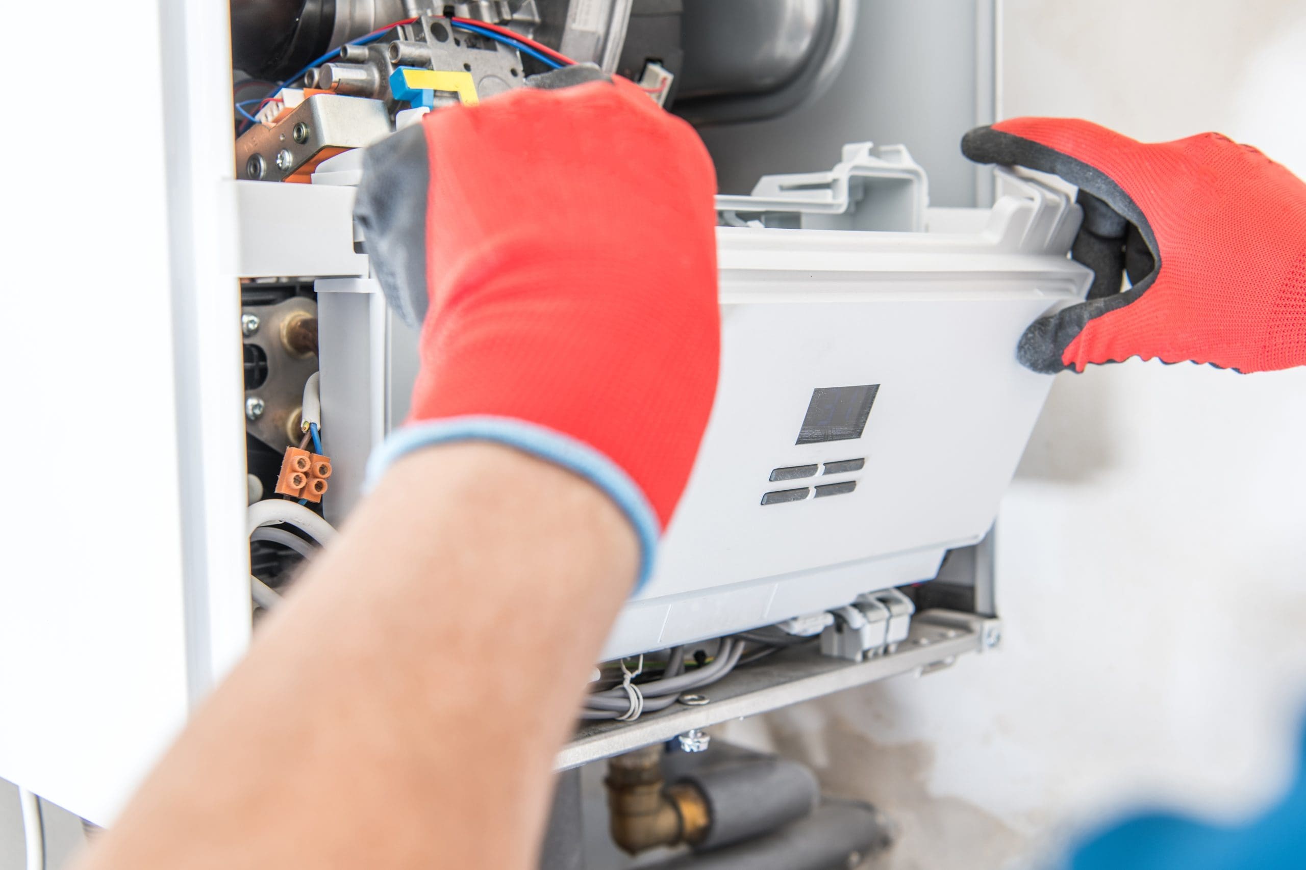 A person wearing red gloves is repairing or installing a boiler. They are handling a panel with electronic components inside the boiler casing. The background is a plain wall.