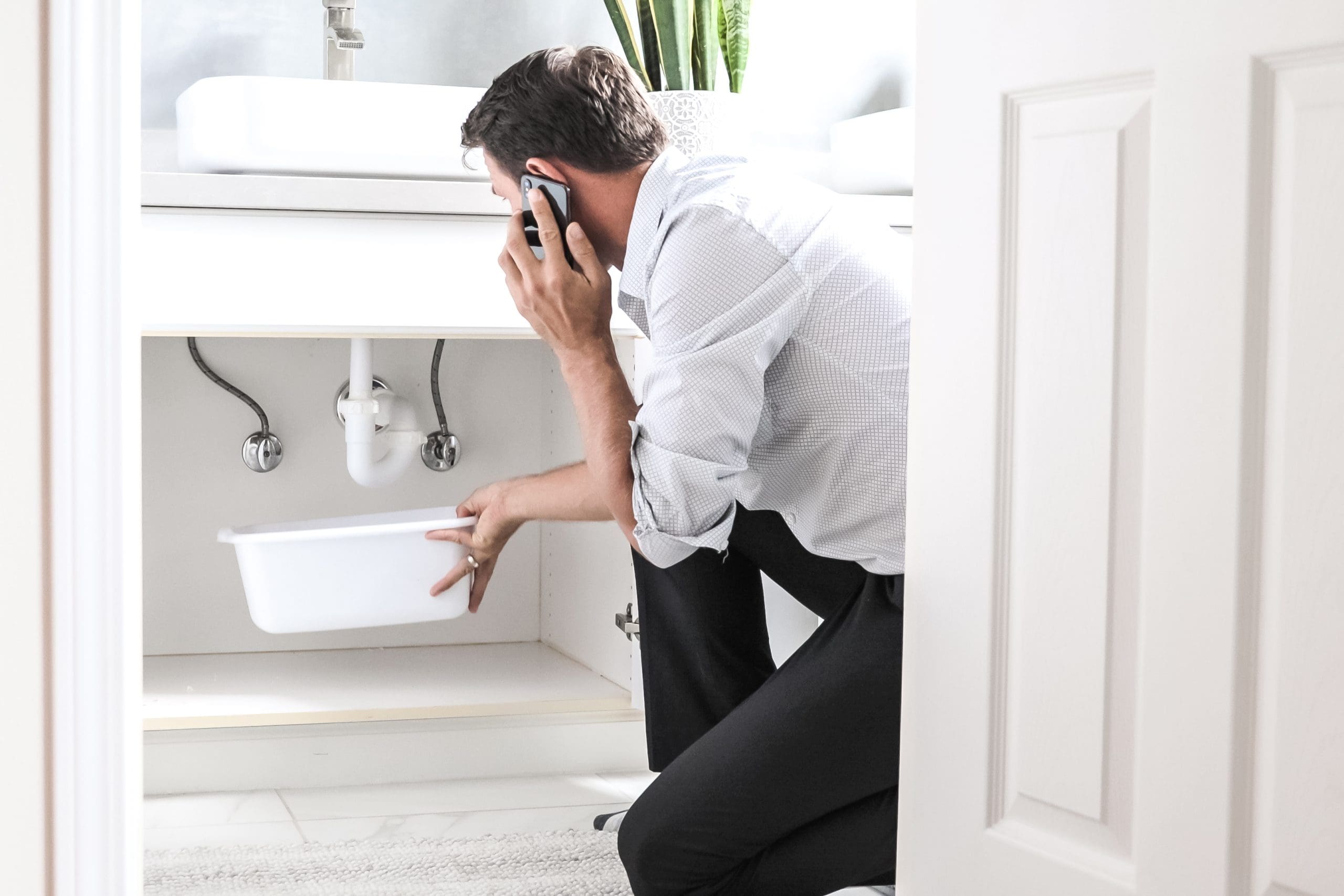 A man in a white shirt is kneeling in front of an open bathroom cabinet. He's holding a phone to his ear and a plastic bin in his other hand, inspecting plumbing under the sink. The room is bright and well-lit.