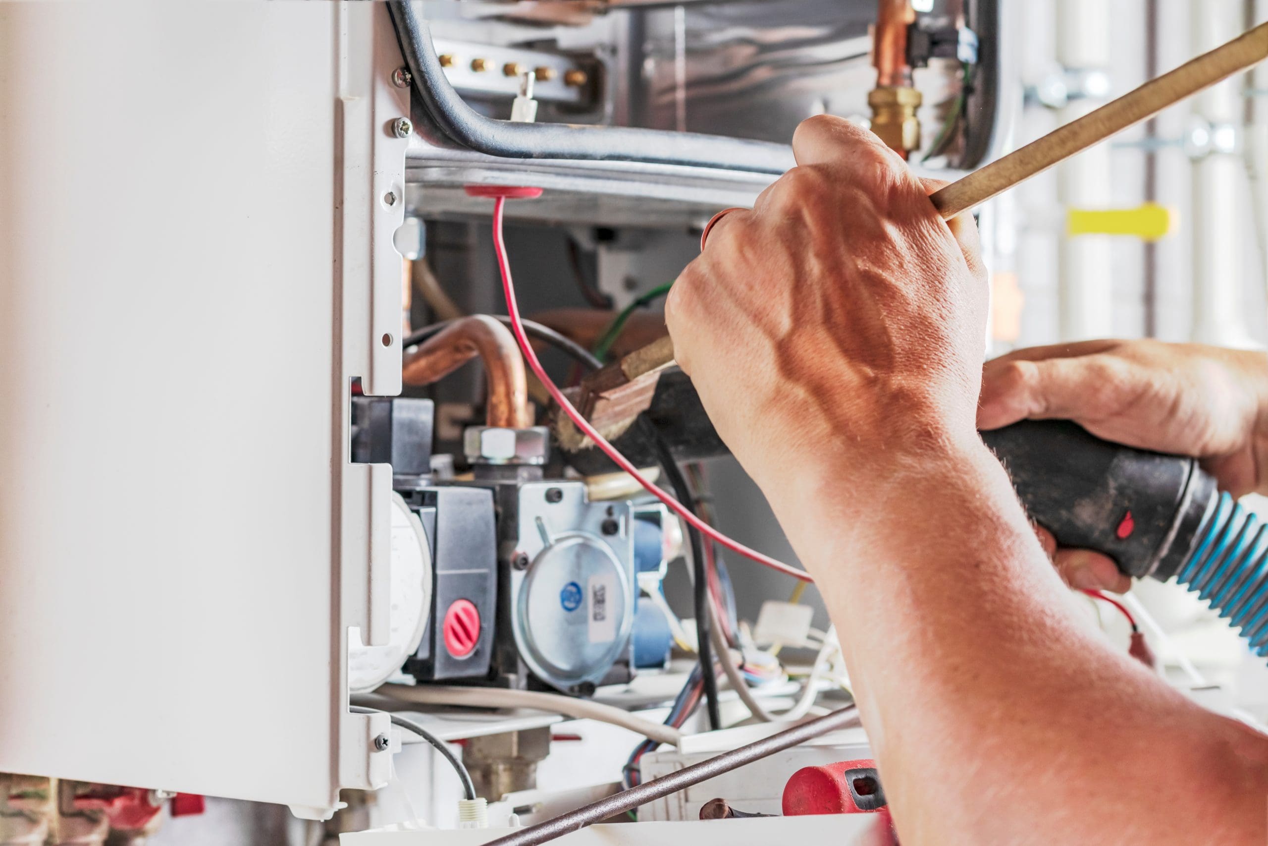 A person working on a gas boiler, using tools to make adjustments. The boiler's interior components, including wires and pipes, are visible, indicating maintenance or repair work.