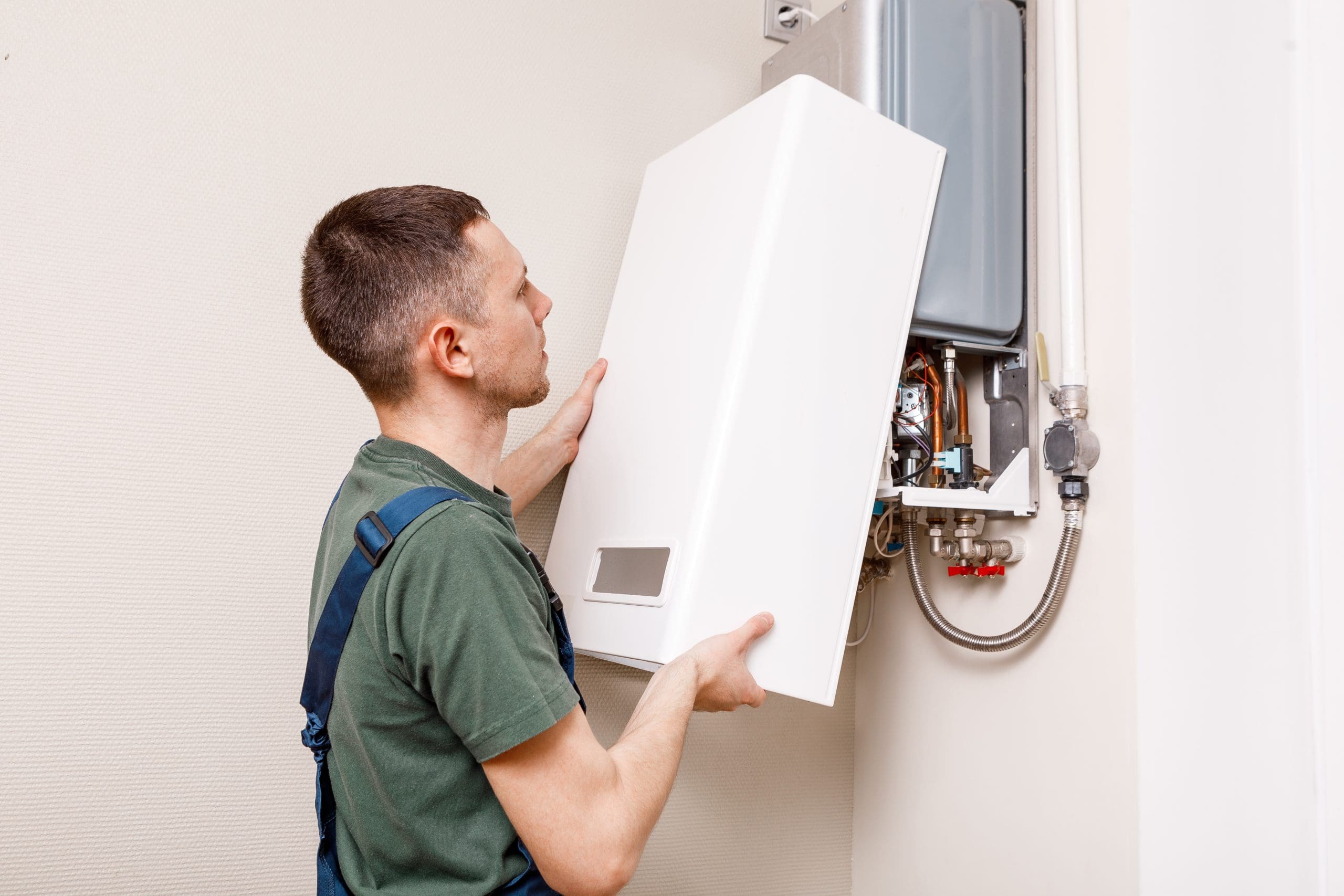 A technician in a green shirt and overalls is examining a wall-mounted boiler. He is holding the front panel, which is open, revealing the internal components of the heating unit. The setting appears to be indoors.