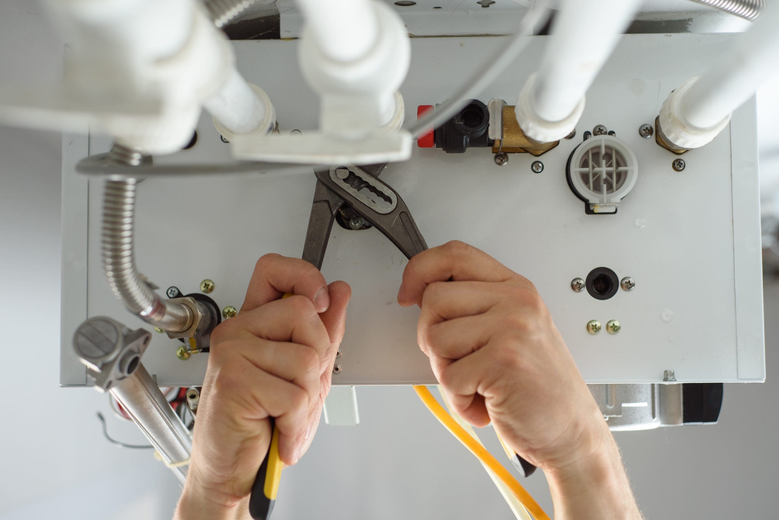 Hands using pliers to adjust pipes on a gas boiler. Several white pipes and metal connectors are visible, along with a yellow tube and various control knobs on the boiler. The photo is taken from underneath the boiler.