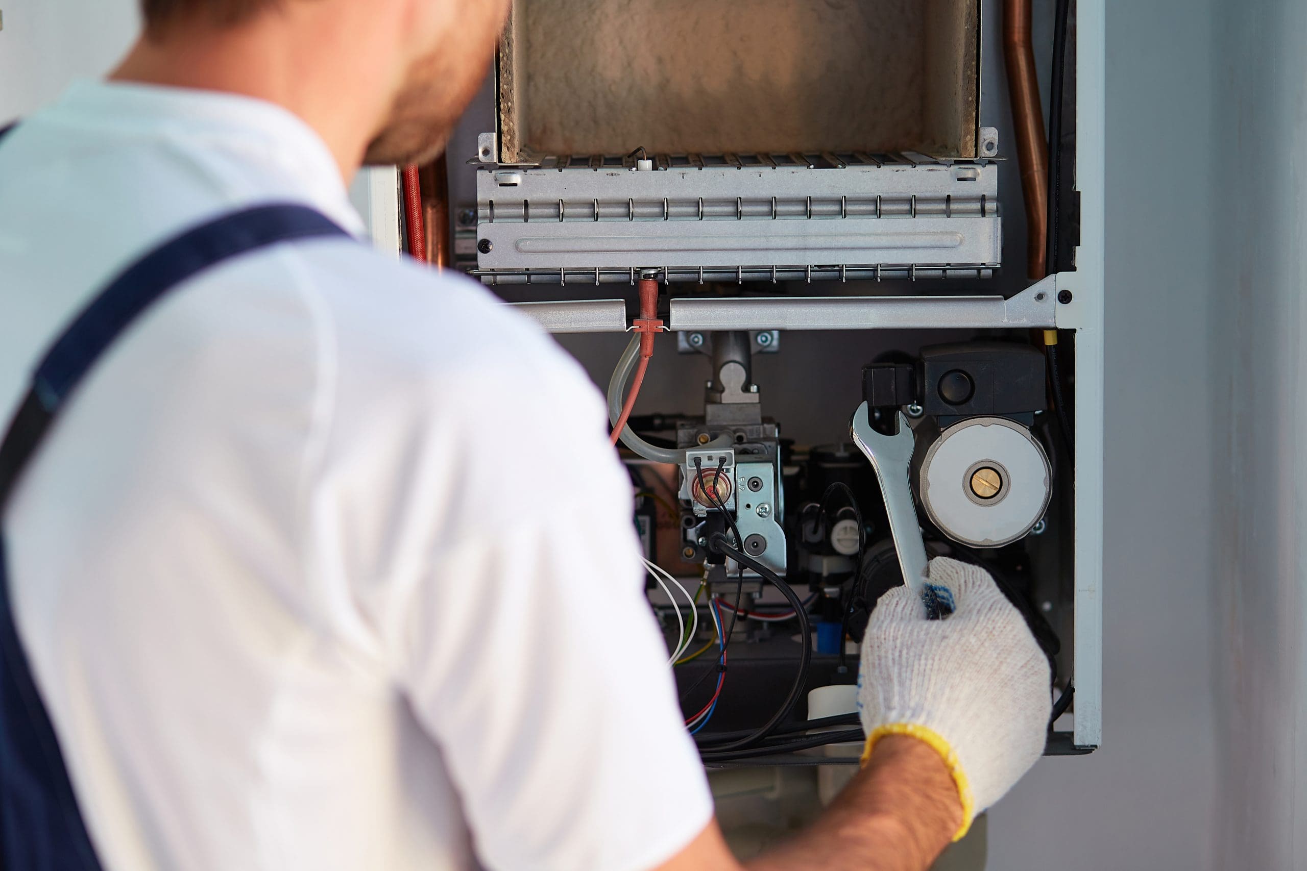 A person in a white shirt and overalls uses a wrench to adjust components inside an open furnace. The person wears a glove on one hand and stands with their back to the camera, focusing on the furnace's mechanical parts.