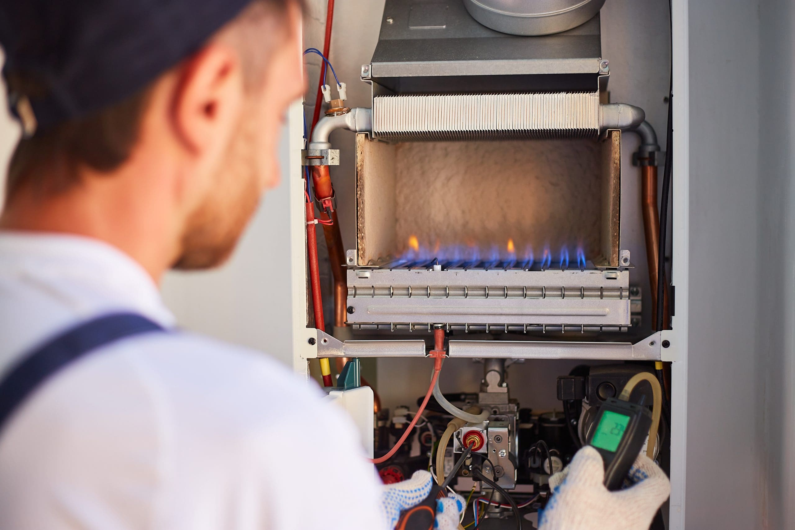 A technician in a cap and gloves is inspecting a gas furnace. The furnace is open, revealing blue flames inside. The technician is holding a device, possibly for measurements or diagnostics.