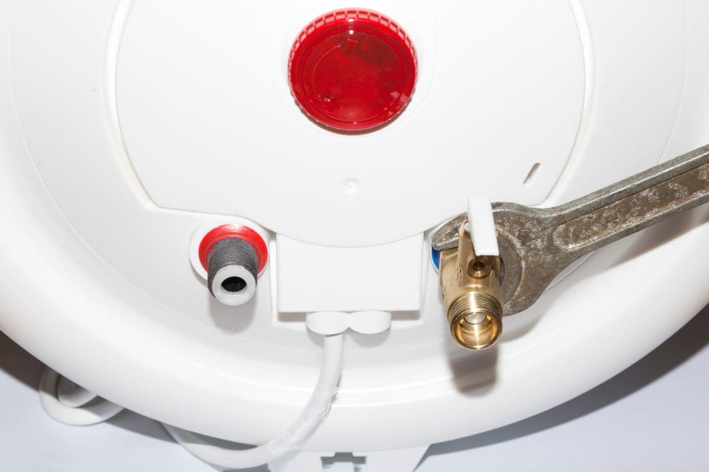 Close-up of a water heater's base showing a wrench turning a brass fitting. The heater is white with a red circular panel and two pipe connections: one red and one black. A white plastic piece is also visible.