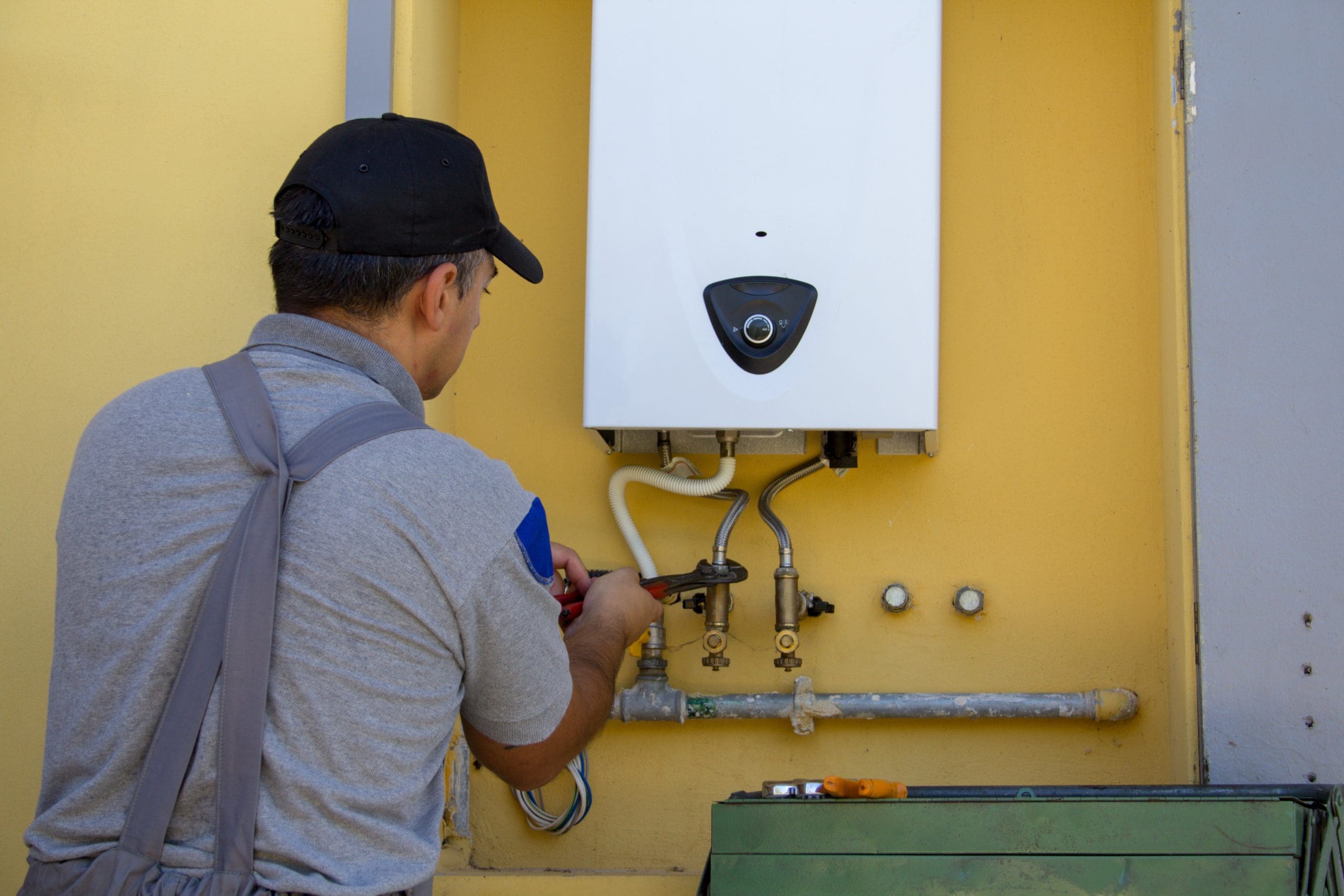 A worker in a gray shirt and black cap is installing or repairing a white water heater mounted on a yellow wall. He is adjusting the pipes with a wrench. Various pipes and valves are visible around the water heater.