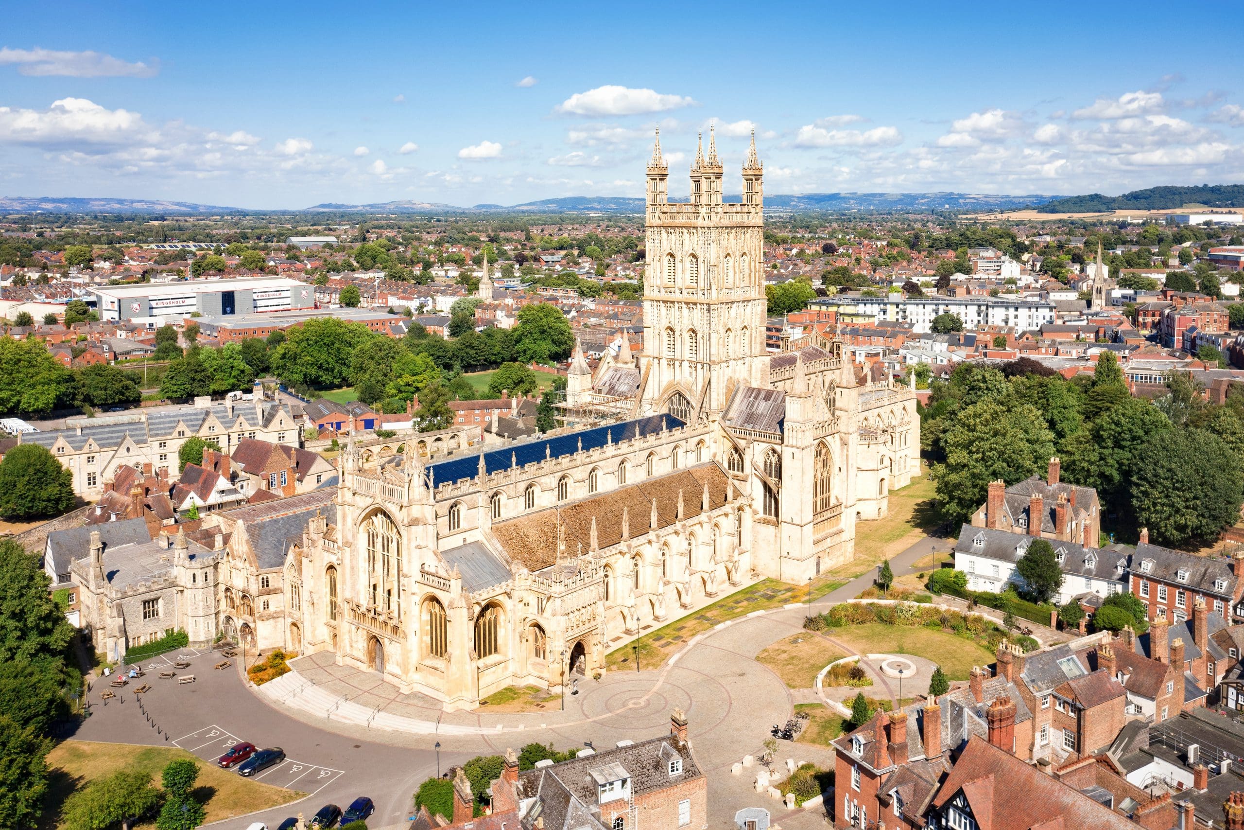 Aerial view of a large, historic cathedral with ornate architecture in a city setting. The building is surrounded by lush greenery and red-roofed houses, set against a backdrop of distant hills under a clear blue sky.