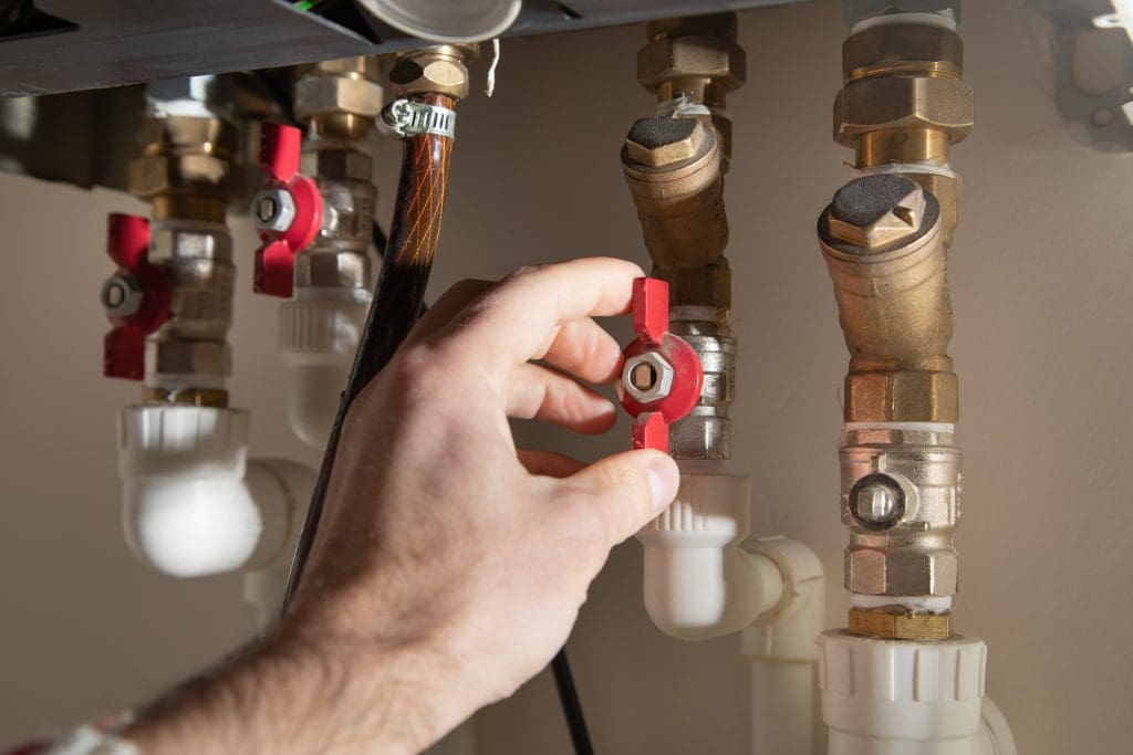 Close-up of a hand adjusting a red valve on a series of brass pipes in a plumbing system. Several pipes and connectors are visible, some with white plastic ends. The scene is well-lit, highlighting the mechanical components.