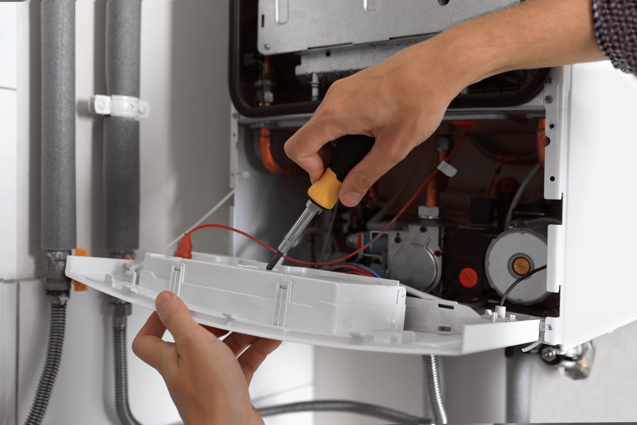 A person uses a screwdriver to repair or maintain a household appliance or machine. Their hands are working on a panel with visible wires and components, focusing on the setup inside a metallic casing.