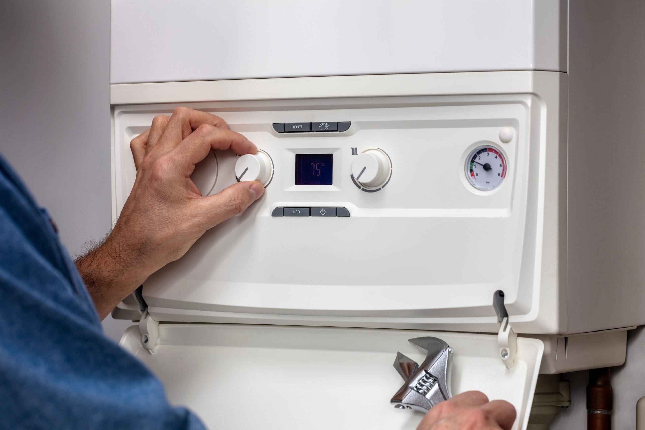 A person adjusts the temperature dial on a white boiler unit, set at 75 degrees. The unit has two knobs and a pressure gauge. A wrench is held in the other hand, indicating maintenance work.