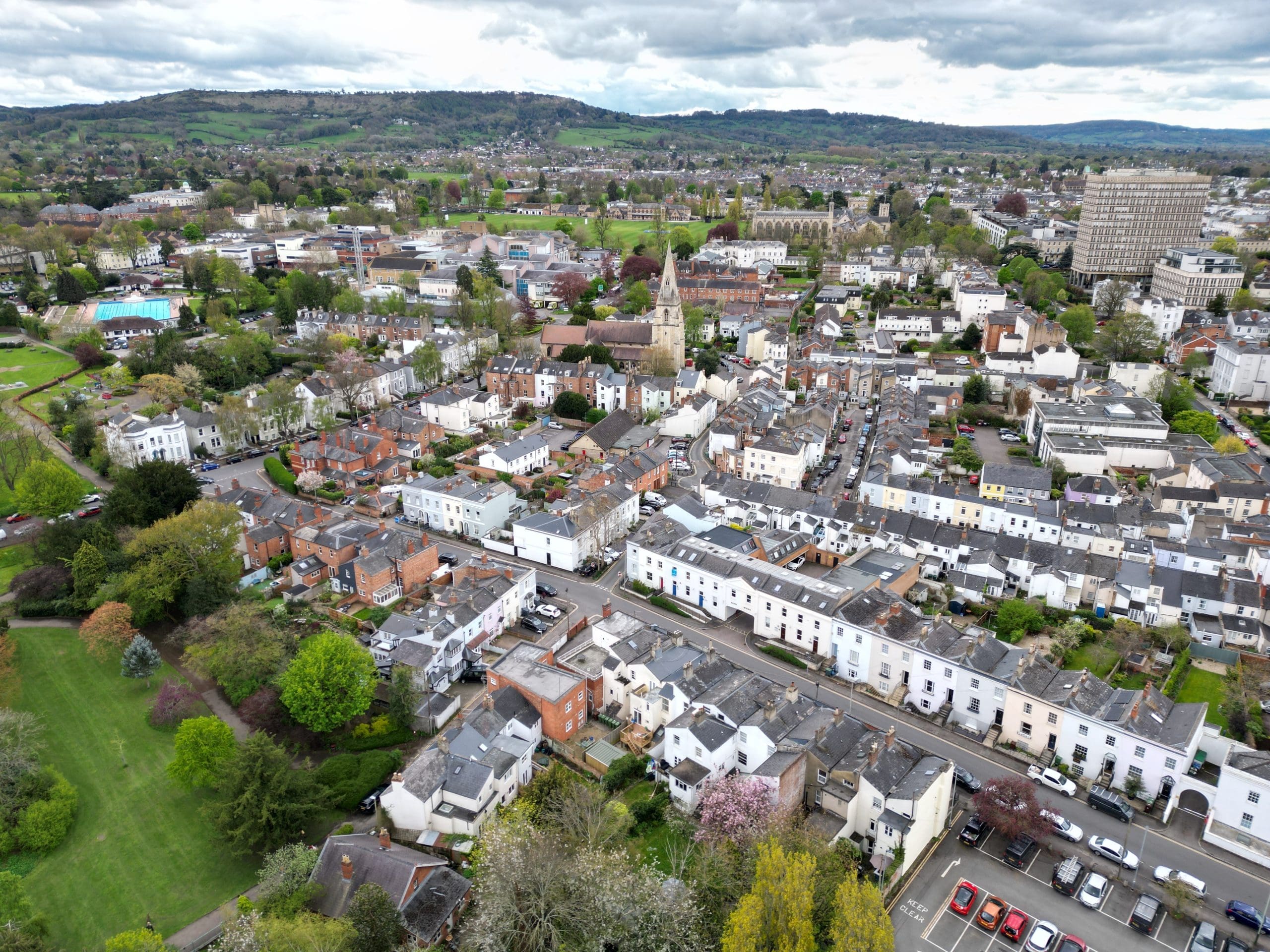 Aerial view of Cheltenham, UK, featuring a mix of historic buildings, residential houses, and lush green parks. Central church spire stands out amidst the townscape, with rolling hills visible in the background under a partly cloudy sky.