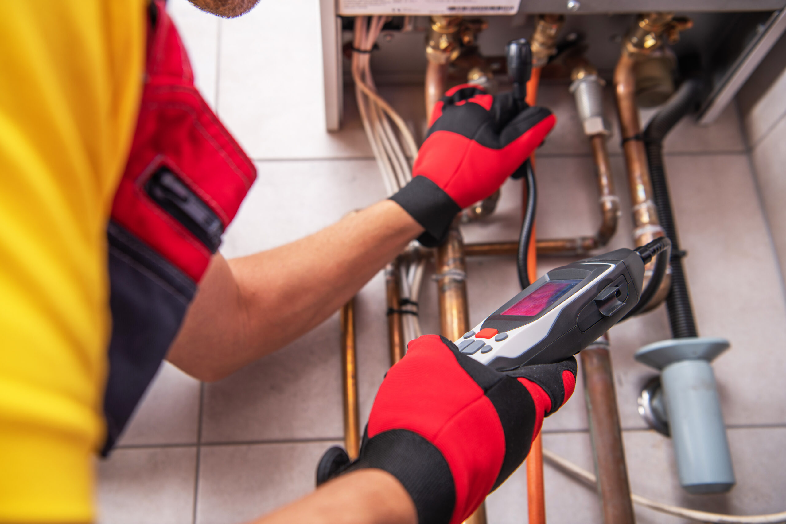 A technician wearing red gloves and a yellow shirt uses a digital diagnostic tool to inspect and measure pipes connected to a boiler or heating system.