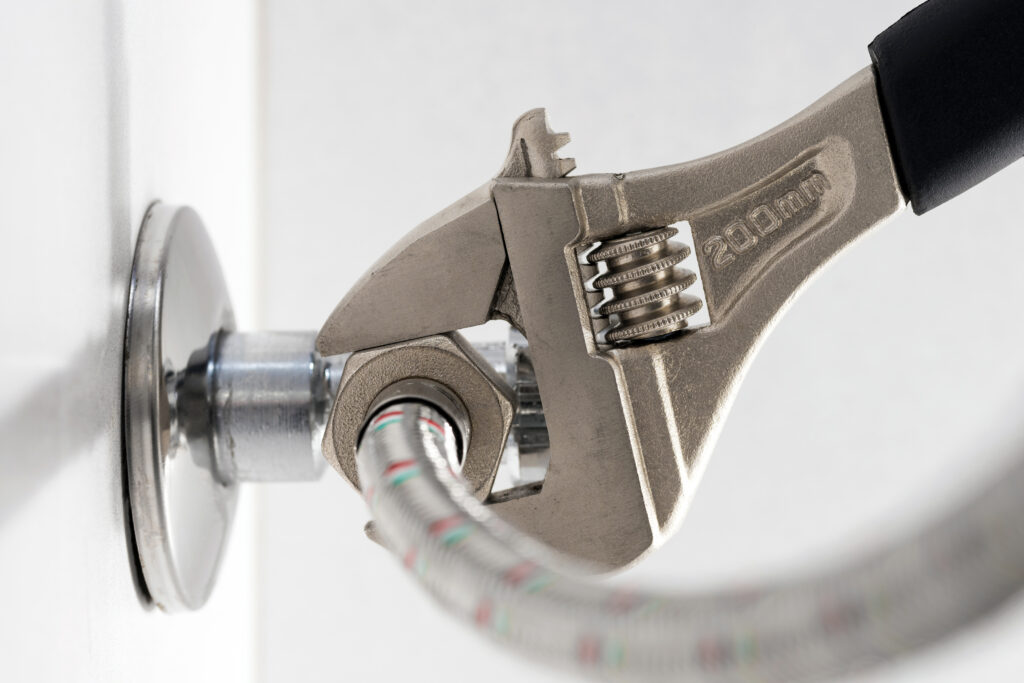An adjustable wrench tightens a metal nut on a flexible hose attached to a wall-mounted water valve.