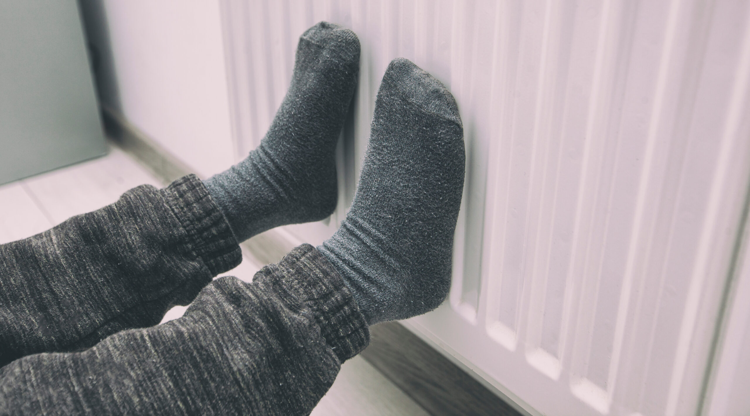 A person wearing gray socks and dark pants sits on the floor, warming their feet against a white radiator in a home setting.