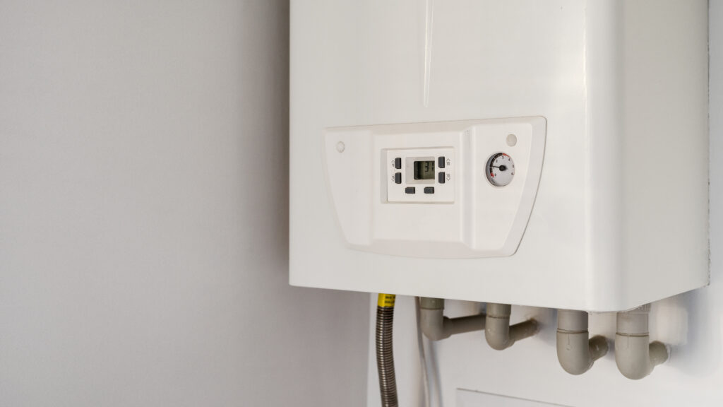 A close-up of a white wall-mounted gas boiler with a digital display and control knobs, connected to several pipes, against a light gray wall.