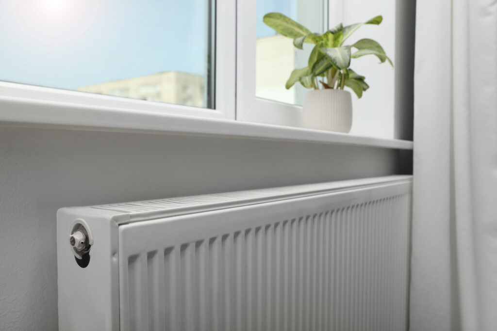 A white radiator is installed beneath a windowsill with a small potted plant on top. Sunlight shines through the window, and a white curtain is partially visible on the right.