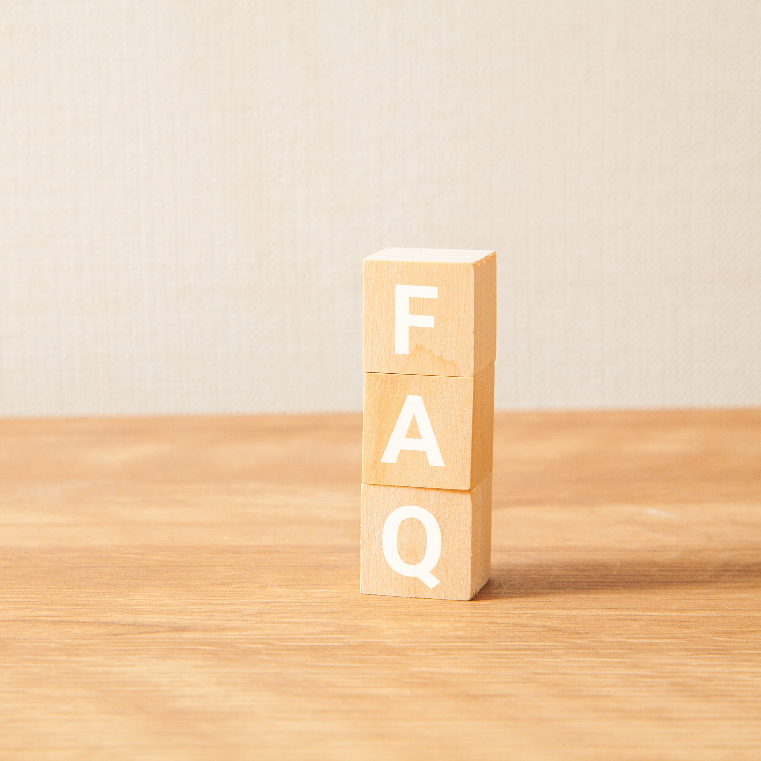 Three wooden blocks stacked vertically on a wooden surface, each with a white letter spelling "FAQ" from top to bottom, against a plain beige background.