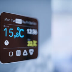 A digital thermostat mounted on a wall displays the room temperature as 15.8°C, the current time as 12:56, and other icons and information on its screen. The background is blurred.
