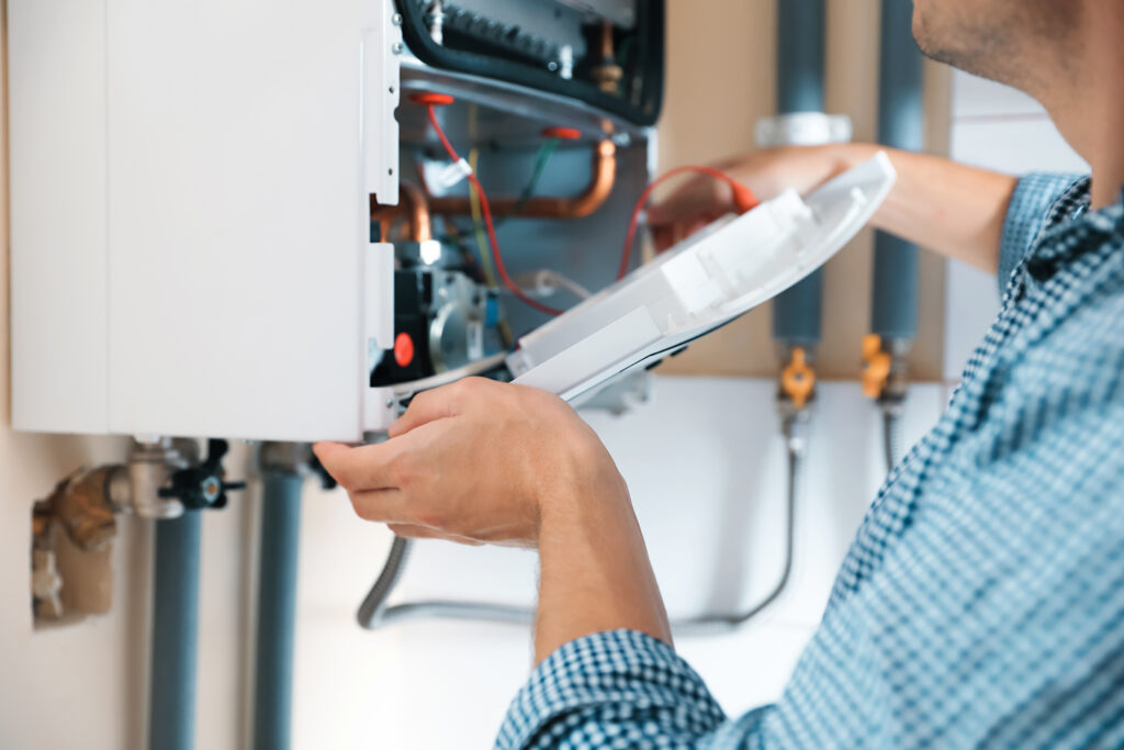 A person in a blue checkered shirt repairs or inspects an open wall-mounted boiler or water heater, exposing wires and internal components. Only the person's arms and torso are visible.