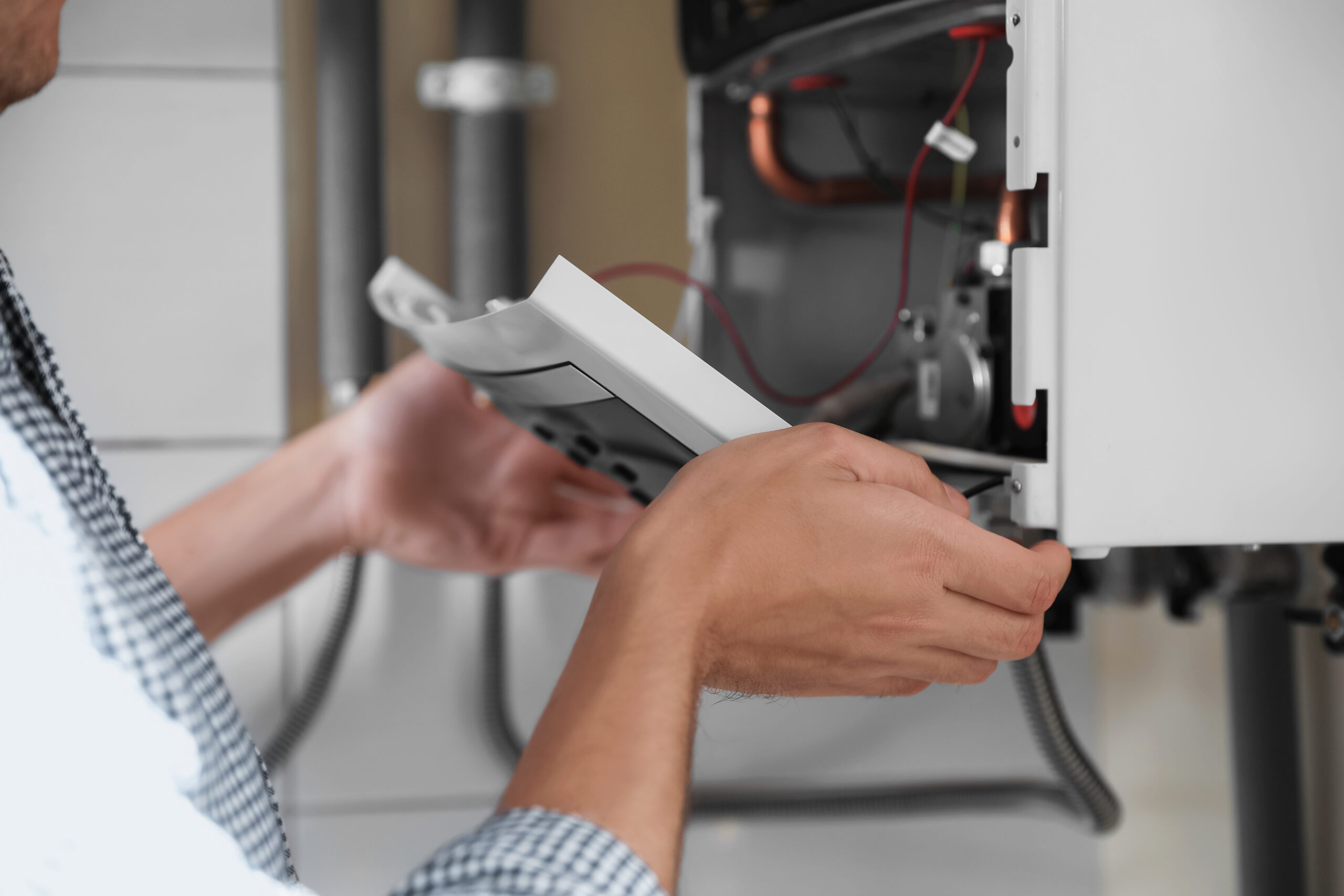 A person in a checkered shirt is repairing or inspecting a wall-mounted boiler, holding and adjusting the front panel with both hands. Exposed wires and internal components are visible.