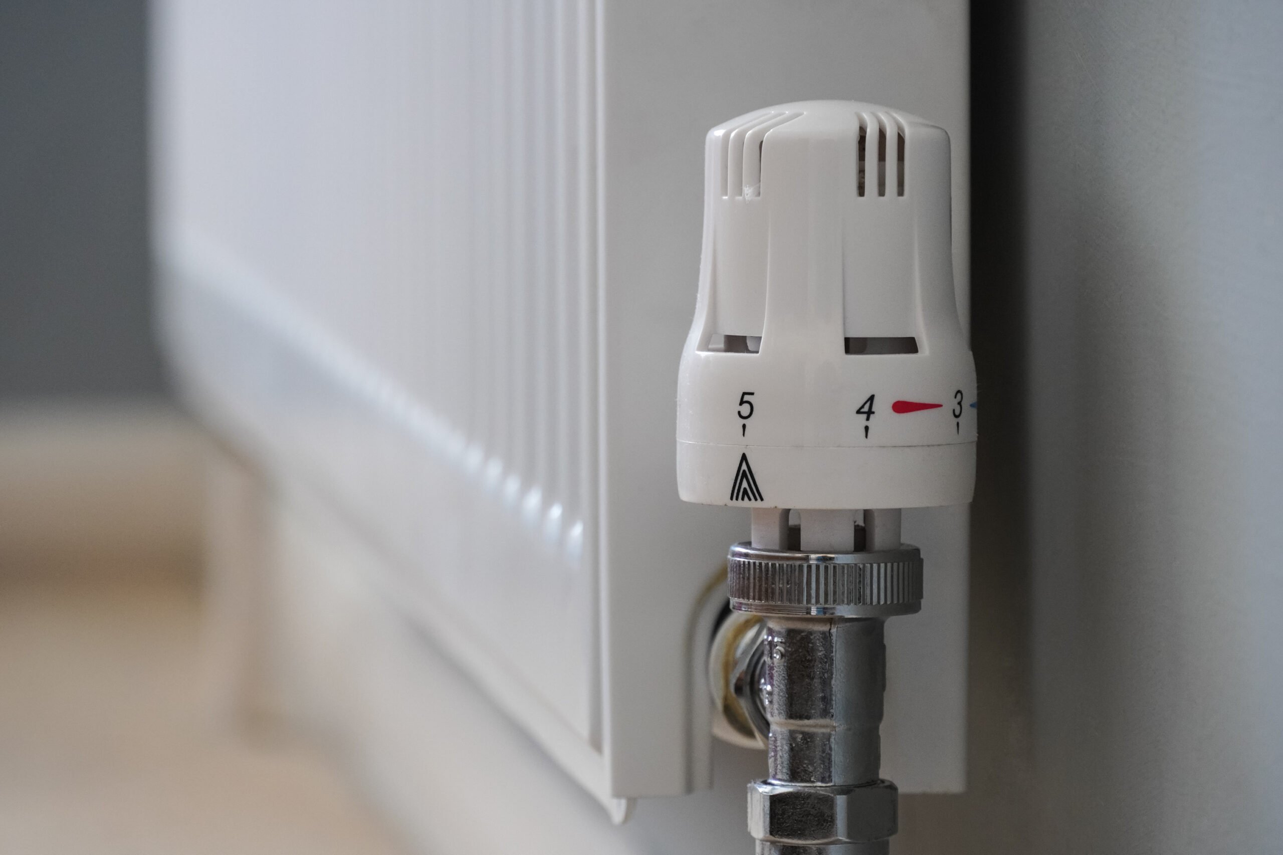 A close-up of a white radiator with a thermostatic valve set between numbers 3 and 4, attached to a silver pipe against a light-colored wall.